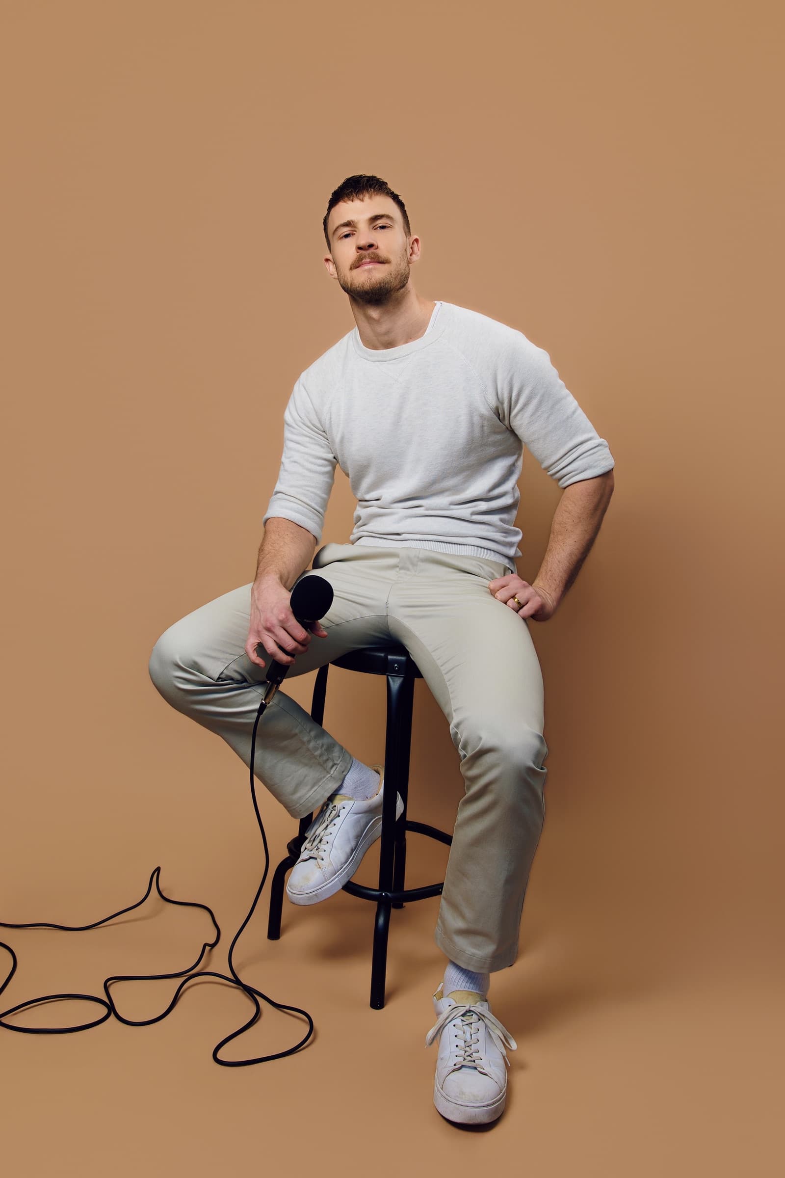 Aidan Wharton seated on a stool with a microphone, photographed against a warm tan studio backdrop.