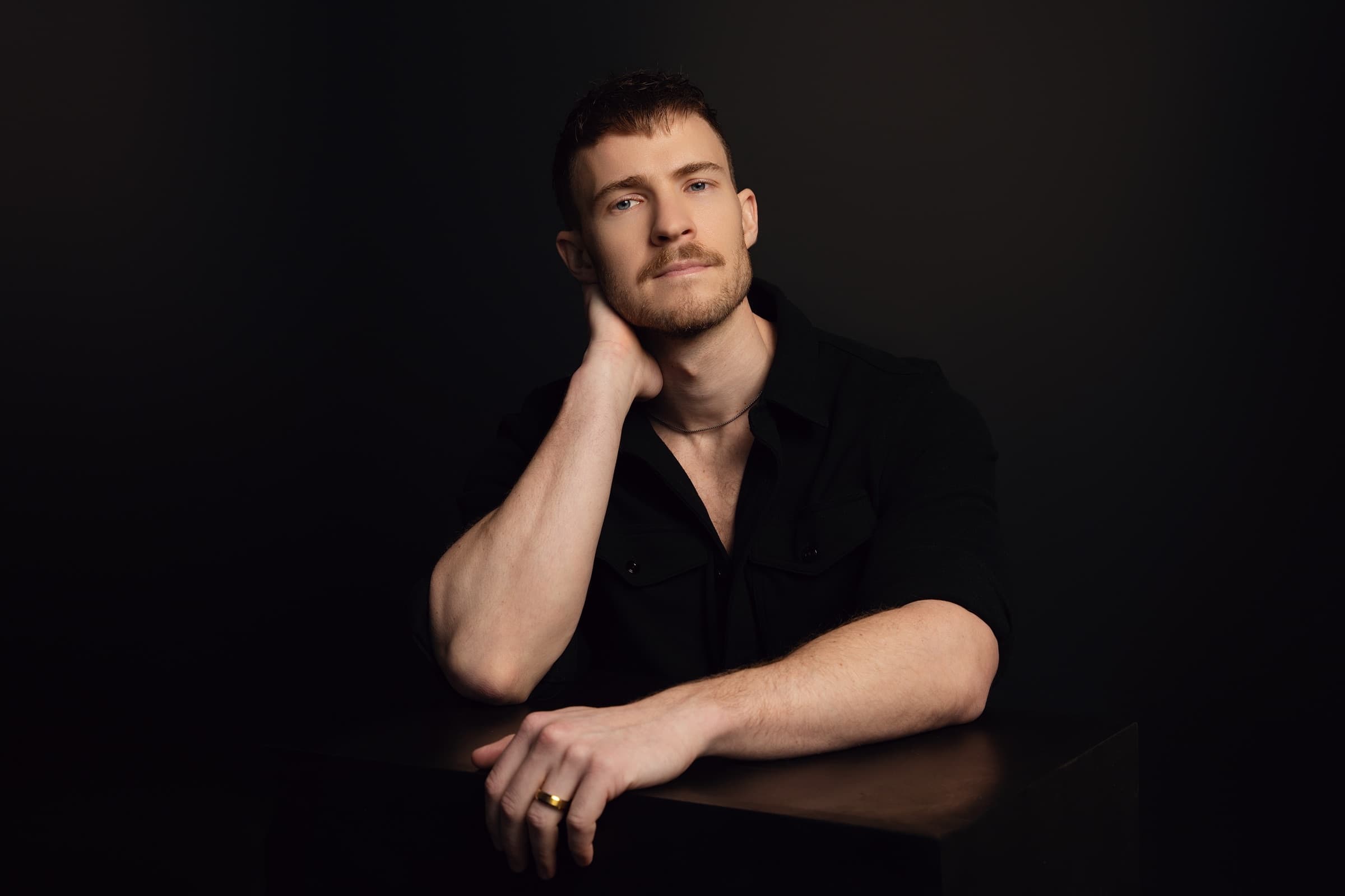 Editorial portrait of Aidan Wharton in a black shirt against a deep black backdrop.